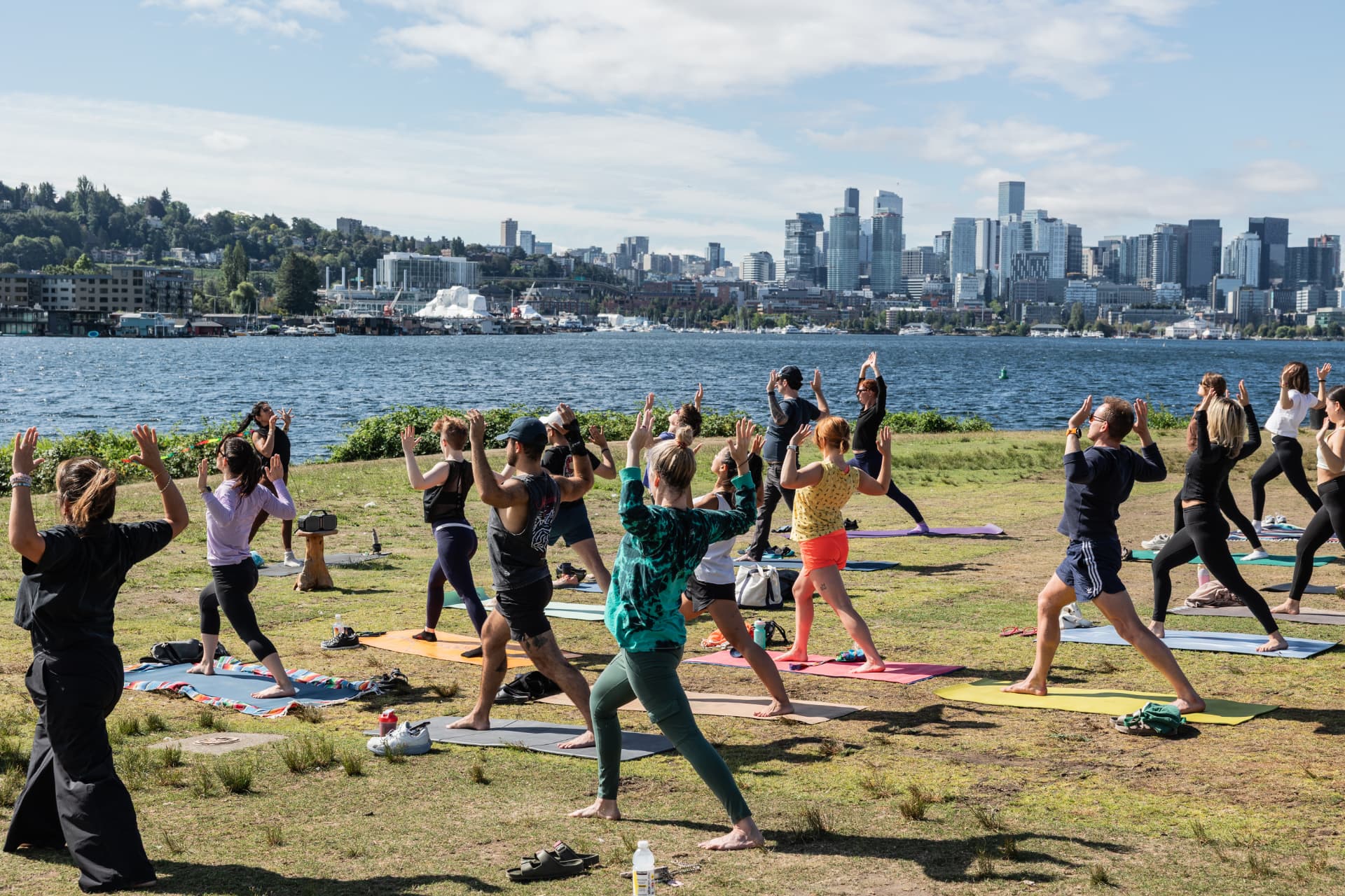 A large outdoor yoga class in warrior pose on the grass at Gas Works Park, with Lake Union and the Seattle skyline behind them.