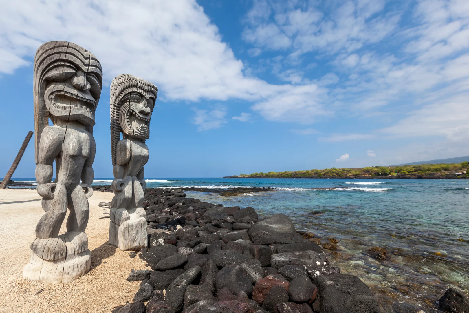 Carved kiʻi standing over the lava rock shoreline and turquoise water of the Kona coast in Hawaiʻi